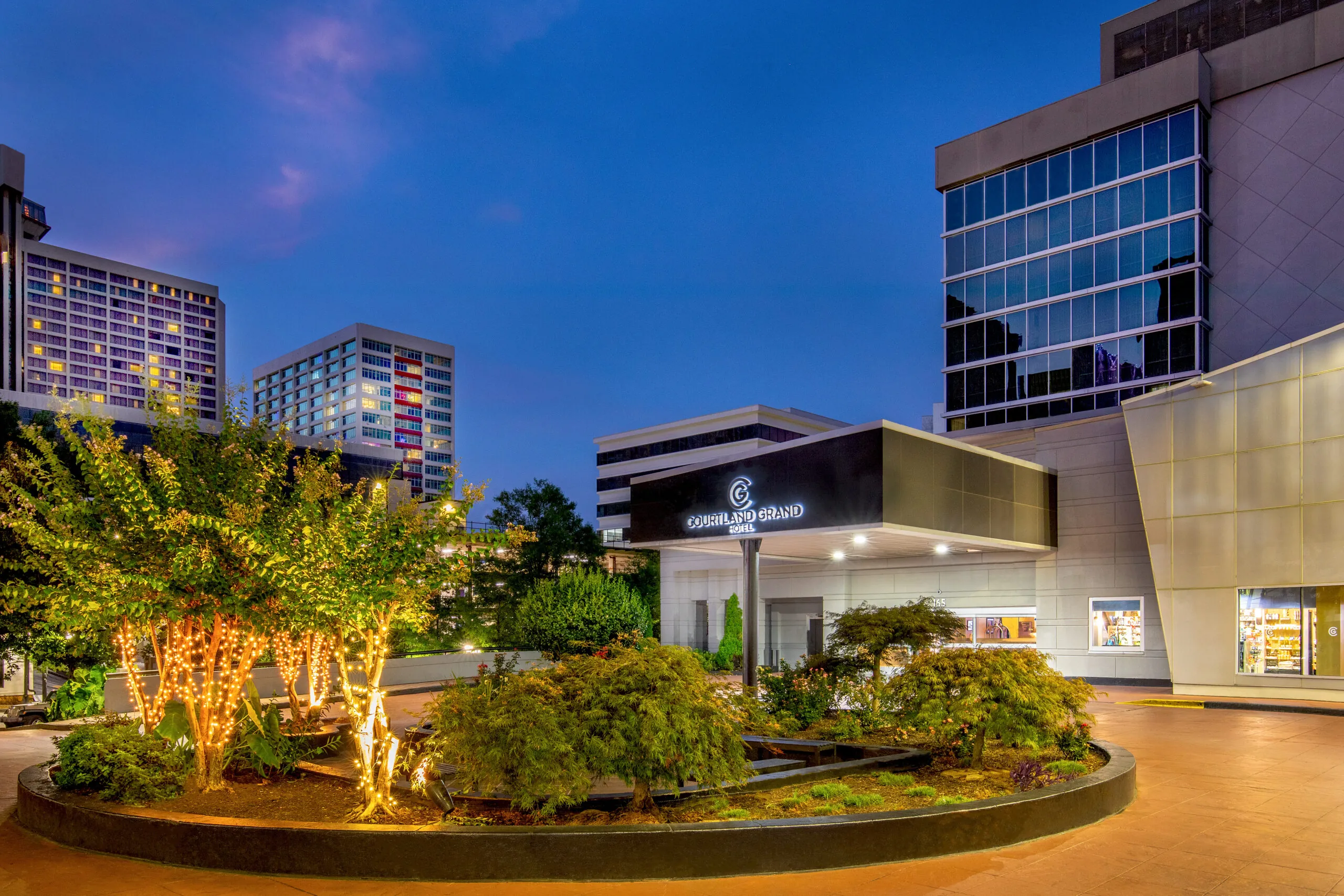 A modern hotel entrance at dusk, featuring the sign “DoubleTree Grand,” surrounded by landscaped greenery and small trees decorated with string lights; tall buildings are visible in the background under a blue-purple sky.